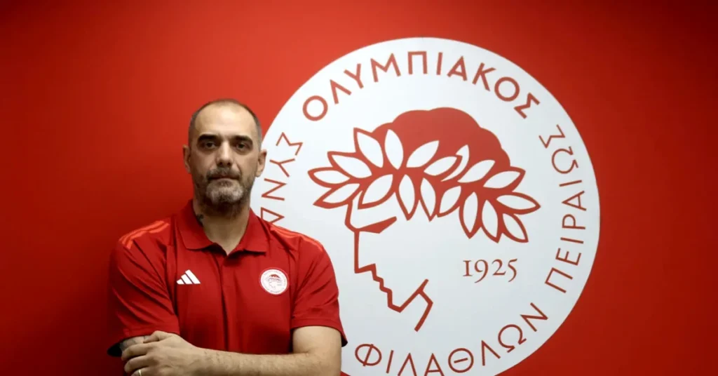 Ο Κώστας Τσιλιμπάρης χαμογελαστός με φόντο το σήμα του Ολυμπιακού μετά την ανάληψη των καθηκόντων του