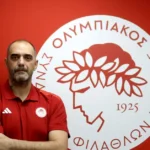 Ο Κώστας Τσιλιμπάρης χαμογελαστός με φόντο το σήμα του Ολυμπιακού μετά την ανάληψη των καθηκόντων του