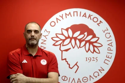 Ο Κώστας Τσιλιμπάρης χαμογελαστός με φόντο το σήμα του Ολυμπιακού μετά την ανάληψη των καθηκόντων του