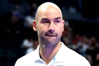 Ο Βασίλης Σπανούλης με κοστούμι, δίνοντας παθιασμένες οδηγίες από τον πάγκο, με φόντο το σήμα του Ολυμπιακού και τις στοιχηματικές αποδόσεις.